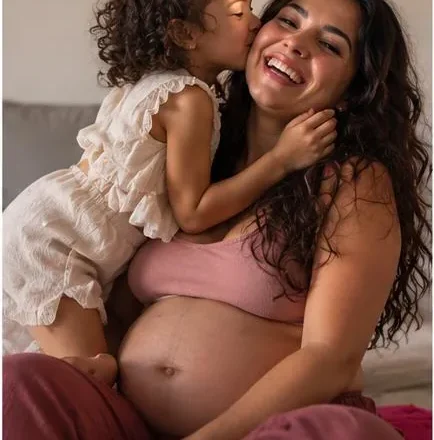 Gestante sorrindo enquanto a filha pequena beija o rosto dela